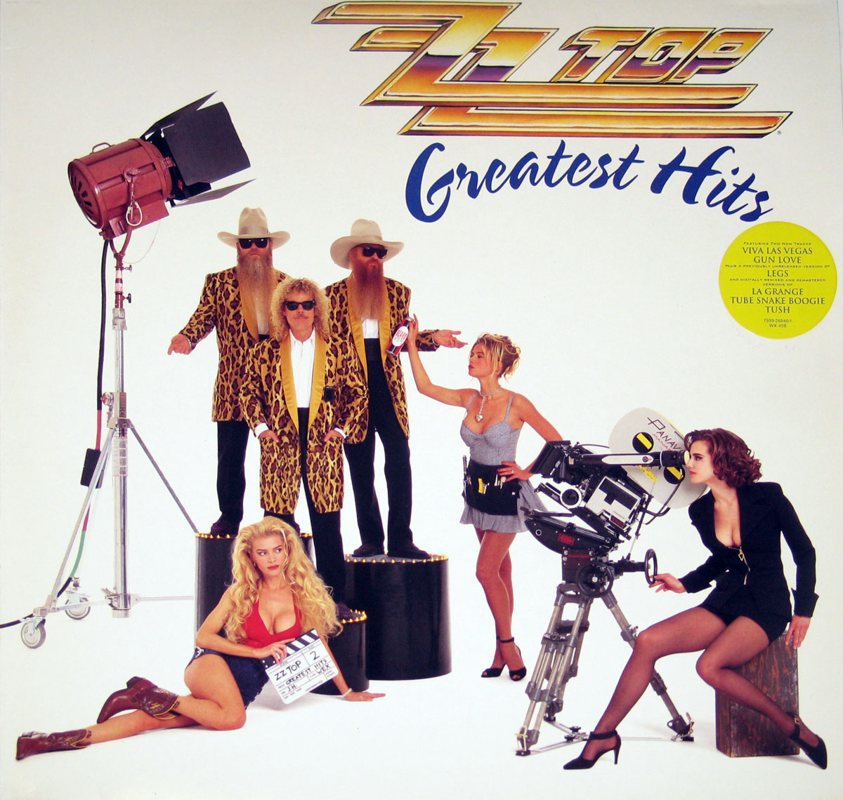 Album cover for ZZ Top’s Greatest Hits, featuring band members Billy Gibbons, Dusty Hill, and Frank Beard in leopard-print jackets and cowboy hats, standing on podiums in a film set. Surrounding them are women dressed in ’90s fashion, one holding a movie clapboard labeled ‘ZZ Top 2,’ while others operate film equipment. The ZZ Top logo and ‘Greatest Hits’ title appear at the top, with a yellow sticker listing songs like ‘Viva Las Vegas,’ ‘Legs,’ and ‘La Grange.’